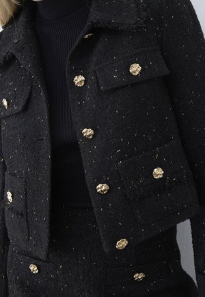 Sort tekstureret jakke med gyldne prikker, med seks udsmykkede gyldne knapper og klaplommer, båret over en sort ribstrikket rullekrave.