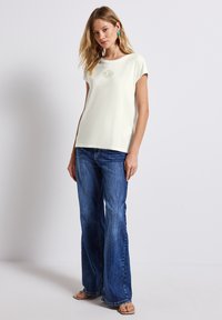Camiseta corta de manga corta color crema con texto bordado, combinada con jeans de mezclilla azul de patas de elefante. Modelo lleva sandalias beige y pendientes grandes multicolores.