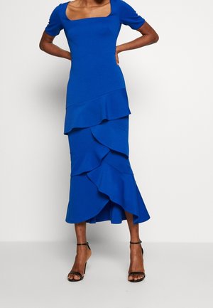 Femme portant une robe midi bleu roi avec une jupe à volants superposés et un décolleté carré, associée à des talons hauts noirs à bride cheville.