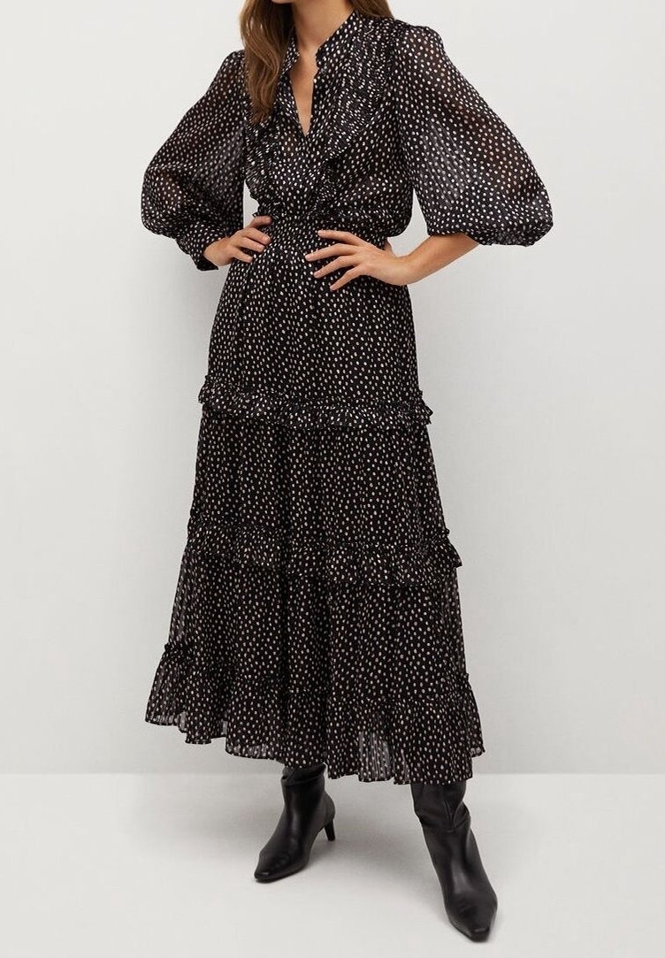Femme portant une robe noire transparente à manches longues avec un petit imprimé floral blanc et des détails à volants, associée à des bottines noires.