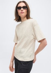 Άτομο με καστανά μαλλιά μήκους μέχρι τους ώμους, που φοράει μαύρα ορθογώνια γυαλιά ηλίου, κρεμ κοντομάνικο πουκάμισο και μαύρο παντελόνι, ποζάροντας μπροστά σε λευκό φόντο.