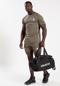 Olijfgroene sportshirt en shorts met witte accenten, met een zwarte sporttas met het "GO WEAR" logo. Sneakers zijn donker met gestructureerde zolen.