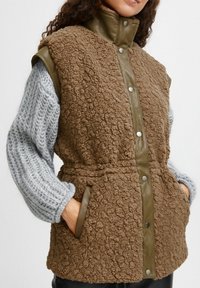 Vesta din fleece maron, cu un guler matlasat de culoare verde măsliniu și nasturi, având buzunare laterale și mâneci tricotate texturate de culoare gri.