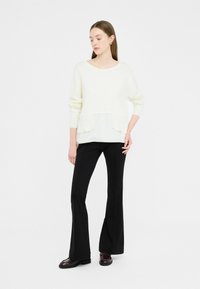 Maglione bianco a maglia con strato testurizzato e due tasche anteriori, abbinato a pantaloni neri a campana e scarpe bordeaux.