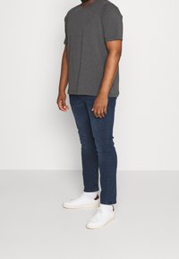Grå crew-neck t-shirt, tætsiddende blå jeans og hvide sneakers med røde detaljer. T-shirten har et lille broderet logo på brystet.