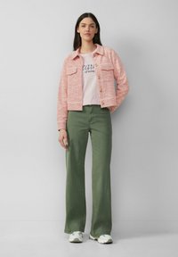 Veste en tweed rose avec deux poches poitrine, assortie à un t-shirt rose clair avec du texte, et un pantalon large vert. Baskets blanches et grises.