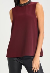 Haut sans manches bourgogne avec un col rond orné de perles noires. Coupe fluide en tissu léger.
