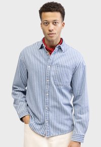 Light blue button-up shirt featuring vertical white stripes, a left breast pocket, and a soft cotton texture, paired with a red bandana.