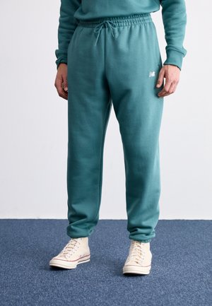 Person iført turkis joggingbukser med snor og beige høje sneakers stående på blåt tæppe.