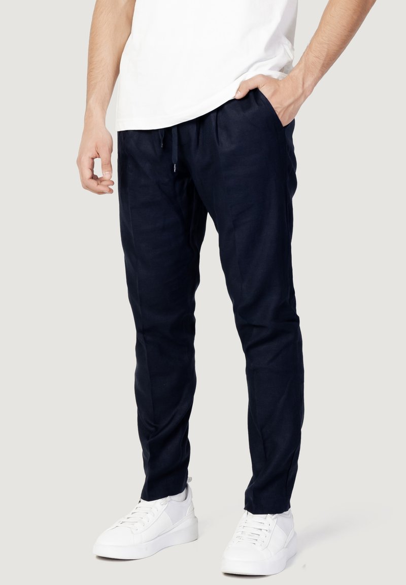 Homme portant un pantalon bleu marine à cordon, des baskets blanches et une chemise blanche à manches courtes, une main dans la poche, debout devant un fond clair.