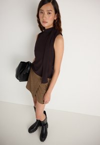 Schwarzes ärmelloses Oberteil mit Drapierungsdetails, brauer karierten Minirock und schwarzen Overkneestiefeln. Schwarze Handtasche mit Knotenverschluss.