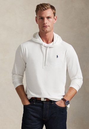 Young man wearing a white hooded sweatshirt with a small logo on the chest, dark jeans, and a wristwatch, standing with hands in pockets.
