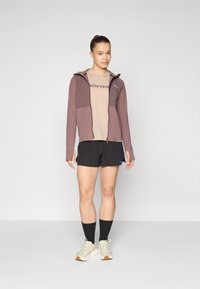 Veste légère à capuche en mauve, associée à une chemise beige et à un short noir. Le modèle porte des chaussures crème et des chaussettes sombres, mettant en avant une tenue sportive.