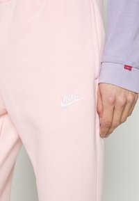 Světle růžové tepláky Nike s bílým vyšívaným logem, s jemnou texturou a uvolněným střihem, spárované se světle fialovým tričkem s dlouhým rukávem.