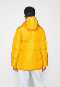 Gul puffjacka med huva, med kviltade sektioner och en glansig yta. Designen inkluderar elastiska ärmslut och en avslappnad passform.