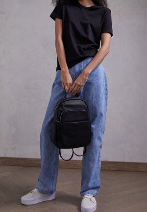 Person wearing black t-shirt, loose blue jeans, and white sneakers holds a small black leather backpack by the top handle.