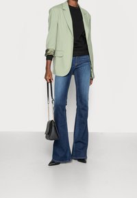 Hellgrüner Blazer, schwarzer strukturierter Pullover, ausgestellte blaue Jeans und schwarze spitz zulaufende Pumps. Kleine schwarze Handtasche mit Kettenriemen.