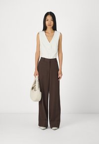 Blouse blanche sans manches avec un col en V, pantalon large marron à taille haute, sac beige et chaussures claires dans un cadre neutre.