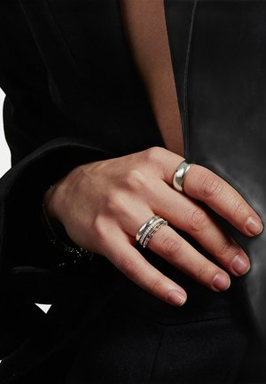 Hånd med fire sølvringe, herunder en simpel ring og tre dekorative ringe, der hviler på sort stof med læderagtig tekstur.