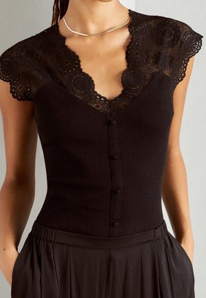 Femme portant un haut noir côtelé avec des manches courtes en dentelle et des détails boutons, associé à un pantalon noir taille haute et un collier fin.