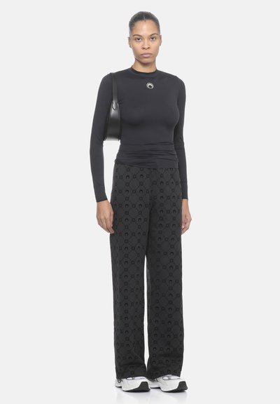 Top nero a maniche lunghe con collo rotondo e logo decorativo, abbinato a pantaloni neri larghi con fantasia e scarpe da ginnastica bianche.