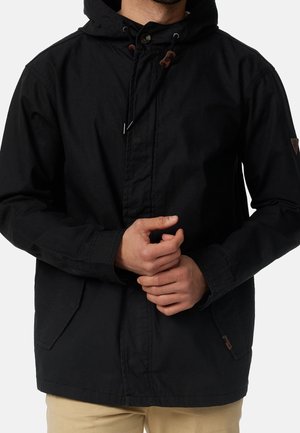 Man met een zwart capuchonjack met voorzakken, die zijn mouw aanpast, beige broek zichtbaar, effen lichte achtergrond.