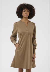 Görbe hajú nő, aki térd alá érő, hosszú ujjú, bézs színű ruhát visel, V-nyakkal és gombbal, keze a zsebében, semleges háttér előtt.