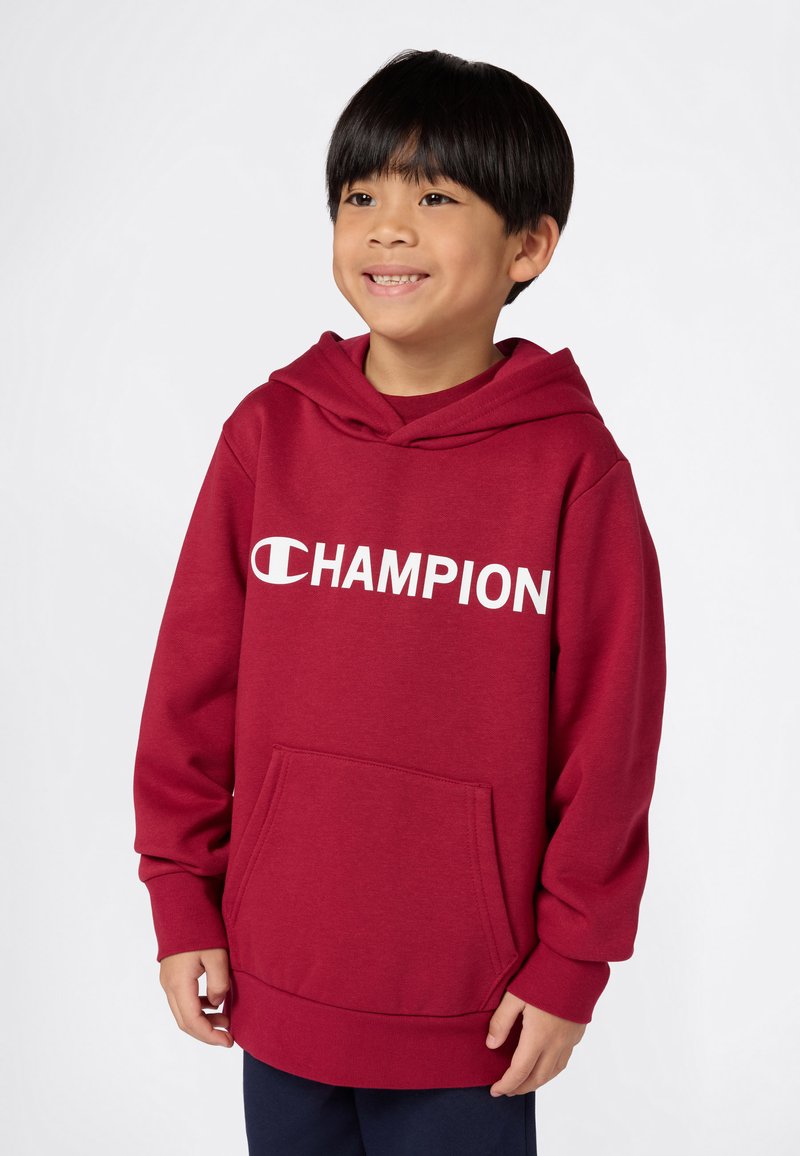 Bambino sorridente con capelli neri che indossa un felpa rossa Champion e pantaloni scuri, in piedi davanti a uno sfondo chiaro e semplice.