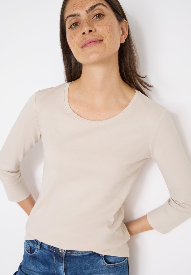 Lys beige langærmet top med rund halsudskæring, lavet af glat stof og med en tæt pasform; parret med blå jeans.