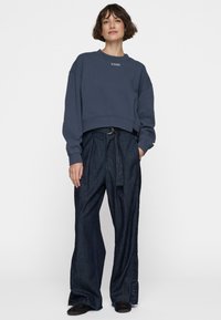 Donna che indossa un felpa corta blu navy con il testo "10DAYS" e pantaloni a gamba larga in denim scuro a vita alta con una cintura, in piedi con una mano in tasca.