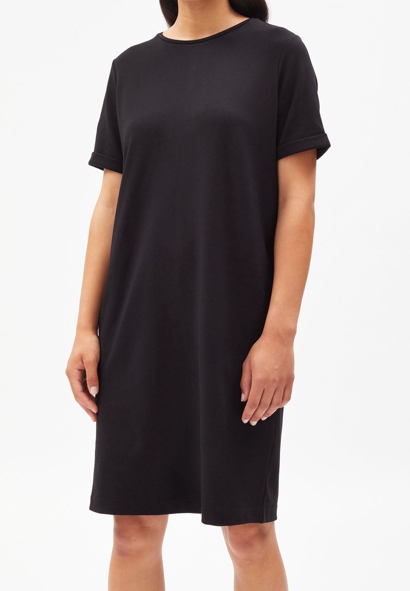 Femme portant une robe noire courte aux manches retroussées arrivant au genou, debout, les bras relâchés le long du corps.