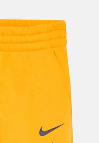 Dzelteni elastīgas jostas šorti ar priekšējo kabatu un melno Nike swoosh logotipu tuvu apakšmalai.