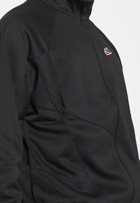 Zwarte ritssluitingjack met hoge kraag, gedragen door een persoon, met een klein rood en wit Nike-logo op de borst.