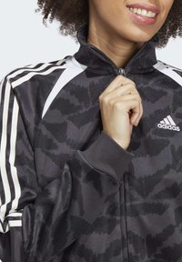 Black jacket with a textured camouflage pattern, white accents, and a zippered collar. Features three white stripes on the sleeves.