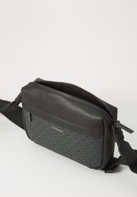 Geantă crossbody neagră cu un capac din piele netedă și un corp din material cu model, având branding gri. Închidere cu fermoar și curea ajustabilă.