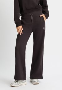 Pantalones de chándal negros de pernera ancha en tejido suave con cintura ajustable mediante cordón y aberturas laterales en el dobladillo. Presenta un pequeño logo en la cadera izquierda.