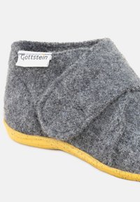 Pantofole di lana grigia con una finitura testurizzata, dotate di una suola in gomma gialla e una cinghia laterale in velcro per una vestibilità regolabile. Etichetta visibile: Gottstein.