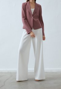 Rosa kavaj med en enda knapp och spetsiga slag, kombinerad med vita vida byxor som har en slät tygstruktur.