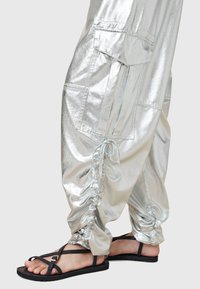 Pantalon argenté brillant froncé avec grande poche latérale, porté avec des sandales plates noires à fines brides et boucle.
