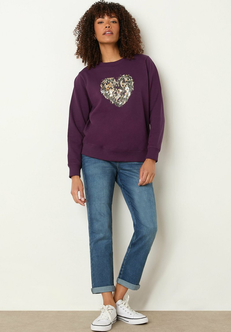 Sudadera morada con un diseño de lentejuelas en forma de corazón en el centro, combinada con jeans de mezclilla remangados y zapatillas blancas. Ajuste casual.