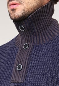 Granatowy sweter dziany z ryżowym kołnierzem, zawierający trzy czarne guziki oraz teksturowany wzór na ciele. Kołnierz wyraźnie się unosi.