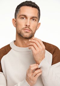 Helle beige Sweatshirt mit gerippter Textur und braunen Wildlederakzenten an den Schultern. Das Modell passt den Ärmel an und zeigt das taillierte Design und die detaillierte Naht.