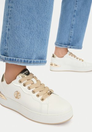 Białe skórzane sneakersy ze złotymi akcentami i beżowymi sznurowadłami, noszone z przyciętymi jasnoniebieskimi dżinsami na białym tle.
