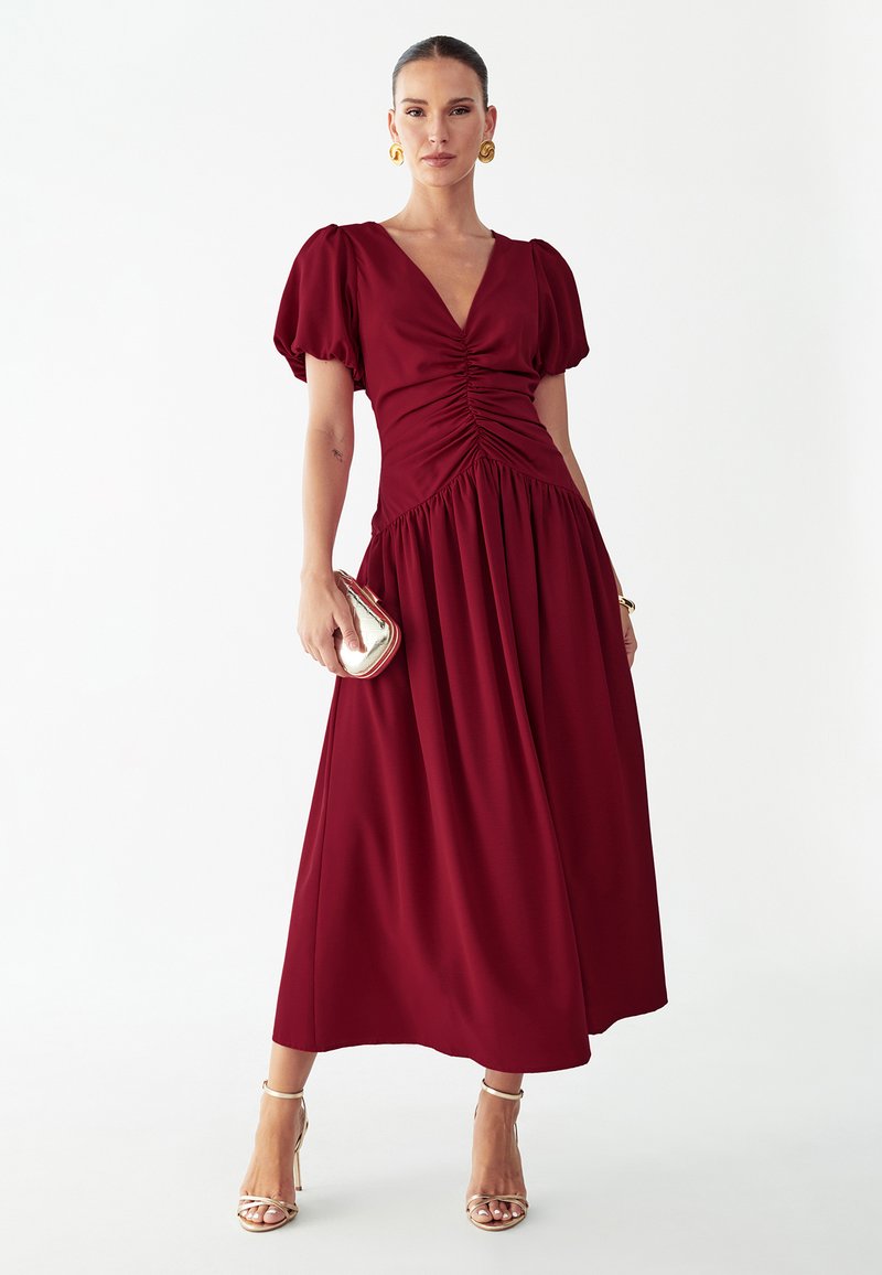 Vrouw die een bordeauxrode midi-jurk met plooien en pofmouwen draagt, gouden hoepeloorbellen, een gouden clutch vasthoudt en gouden sandalen met hoge hakken draagt.