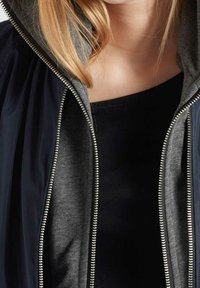Hoodiejacka med grå innerfoder och en glänsande svart ytterlager. Har en lång dragkedja och kontrasterande texturer.