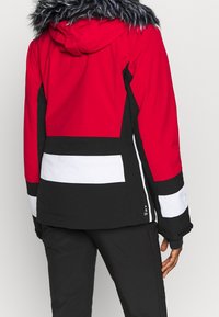 Persona vistiendo una chaqueta de invierno roja, negra y blanca con capucha forrada de piel, mostrada de espaldas contra un fondo liso.