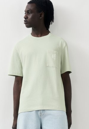 Άνδρας με σκοτεινό δέρμα και κοντά dreadlocks, που φοράει ανοιχτό πράσινο κοντομάνικο μπλουζάκι με τσέπη στο στήθος και ανοιχτό μπλε τζιν.