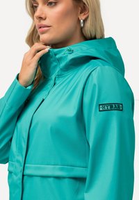 Teal vattentät jacka med huva, med en framsida som har dragkedja, justerbara snoddar och en svart logotyp på ärmen. Slät yta.