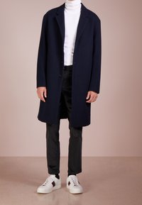 Marineblauer Wollmantel mit geradem Schnitt, einreihigem Design, getragen über einem weißen Rollkragenpullover und schwarzer Hose, kombiniert mit weißen Sneakern.