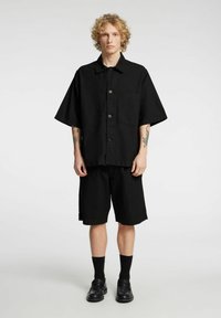 Camisa negra de manga corta con botones y un bolsillo, combinada con shorts negros a juego. La tela parece ligera y con textura.
